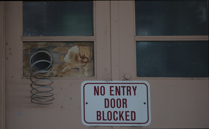 "No entry door blocked" sign on door with rotting wood in place of a window and failing hose duct sticking through